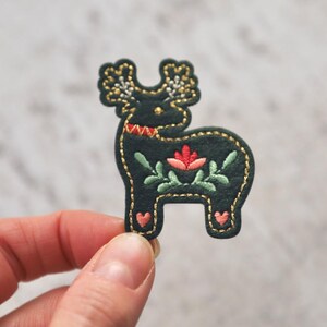 Peut inclure: Un renne en feutre vert fonc&eacute; avec des coutures dor&eacute;es et des d&eacute;tails brod&eacute;s, dont une fleur rose et des feuilles vertes.