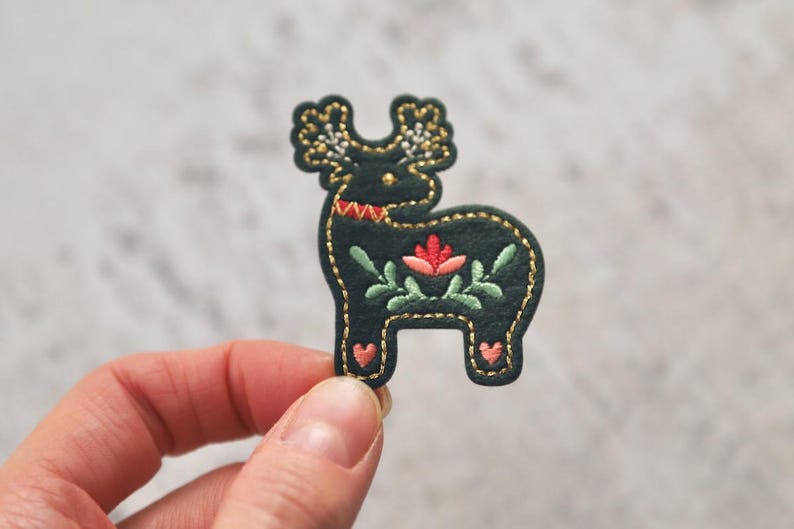 Peut inclure: Un renne en feutre vert fonc&eacute; avec des coutures dor&eacute;es et des d&eacute;tails brod&eacute;s, dont une fleur rose et des feuilles vertes.