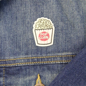 May include: A white and red embroidered iron-on patch of a popcorn bucket with the text "POP CORN" on a blue denim jacket.