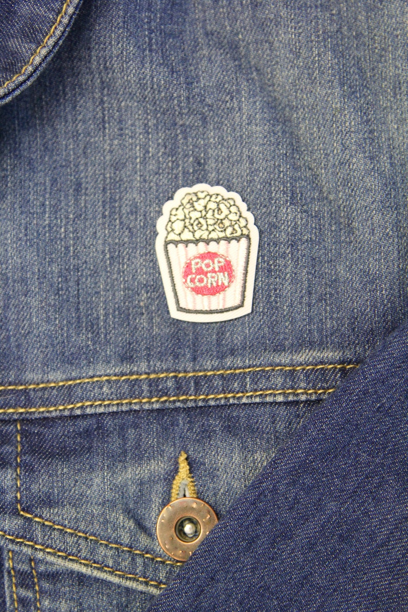 May include: A white and red embroidered iron-on patch of a popcorn bucket with the text "POP CORN" on a blue denim jacket.