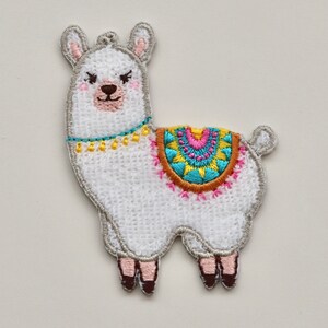 Puede incluir: Aplique de llama blanca bordada con un dise&ntilde;o geom&eacute;trico colorido en su espalda. La llama tiene una nariz marr&oacute;n y ojos negros.