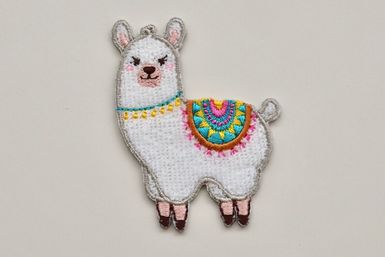 Puede incluir: Aplique de llama blanca bordada con un dise&ntilde;o geom&eacute;trico colorido en su espalda. La llama tiene una nariz marr&oacute;n y ojos negros.