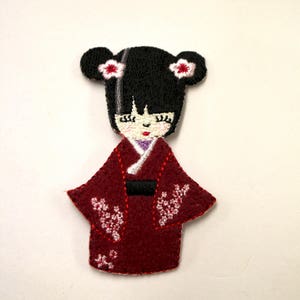 May include: Embroidered felt patch of a Japanese woman wearing a red kimono with white floral designs. She has black hair with white flowers on each side of her head.