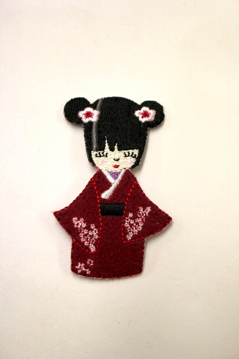 May include: Embroidered felt patch of a Japanese woman wearing a red kimono with white floral designs. She has black hair with white flowers on each side of her head.