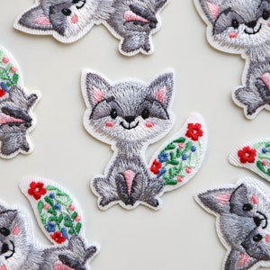 May include: A set of nine embroidered iron-on patches featuring gray foxes with floral tails. The foxes have pink noses and white accents on their faces. The flowers are red, blue, and green.