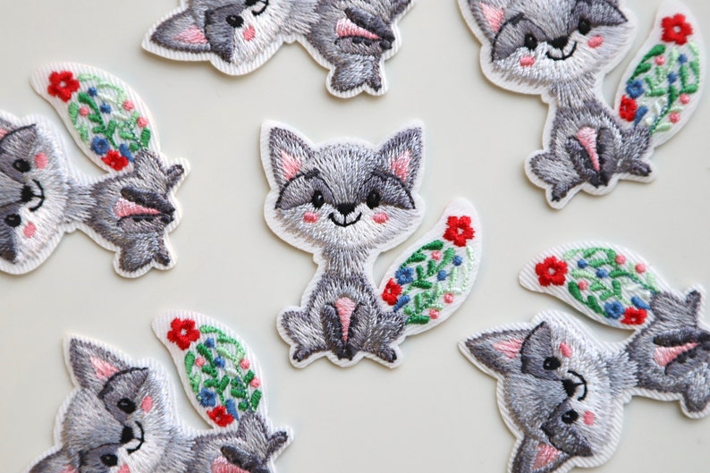 May include: A set of nine embroidered iron-on patches featuring gray foxes with floral tails. The foxes have pink noses and white accents on their faces. The flowers are red, blue, and green.