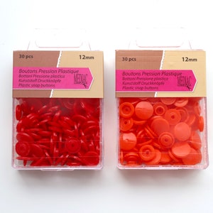 K&ouml;nnte beinhalten: Zwei Packungen mit je 30 roten und orangen Kunststoff-Druckkn&ouml;pfen, jeweils 12 mm im Durchmesser. Die Packungen sind mit "Boutons Pression Plastique" und "MEDIAC" beschriftet.