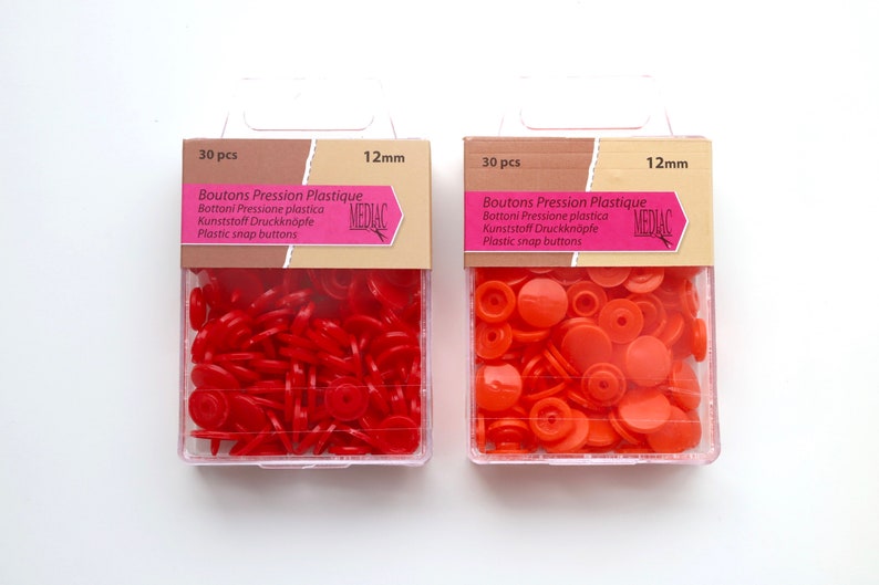 K&ouml;nnte beinhalten: Zwei Packungen mit je 30 roten und orangen Kunststoff-Druckkn&ouml;pfen, jeweils 12 mm im Durchmesser. Die Packungen sind mit "Boutons Pression Plastique" und "MEDIAC" beschriftet.