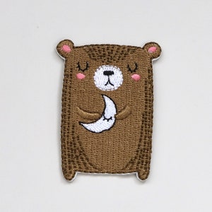 May include: Embroidered brown bear patch with closed eyes, holding a white crescent moon. The bear has pink cheeks and a white nose.