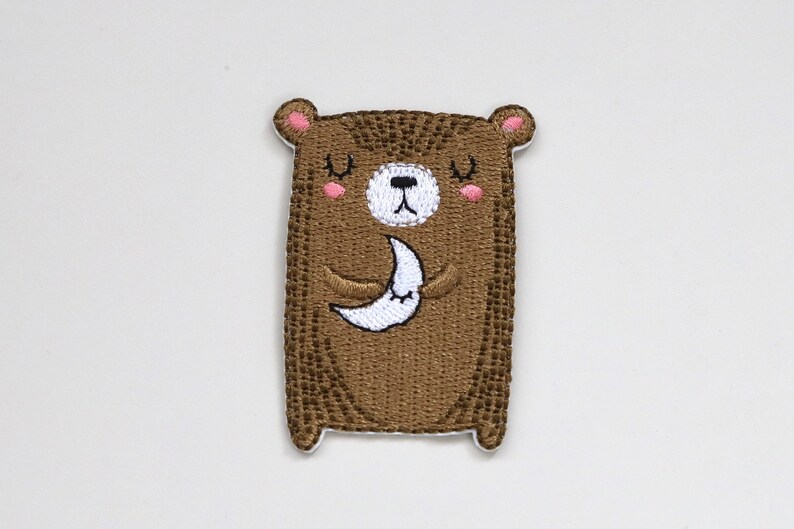 May include: Embroidered brown bear patch with closed eyes, holding a white crescent moon. The bear has pink cheeks and a white nose.