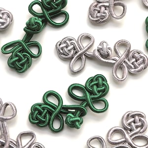 May include: A collection of green and silver Chinese knot toggles, also known as frog closures. These decorative fasteners are used for clothing, bags, and other crafts.