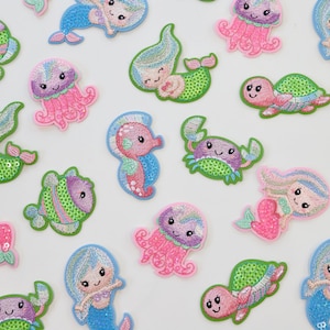 May include: A collection of colourful, embroidered iron-on patches featuring various sea creatures, including mermaids, jellyfish, seahorses, crabs, and turtles. The patches are made with sequins and have a whimsical, cartoon-like style.