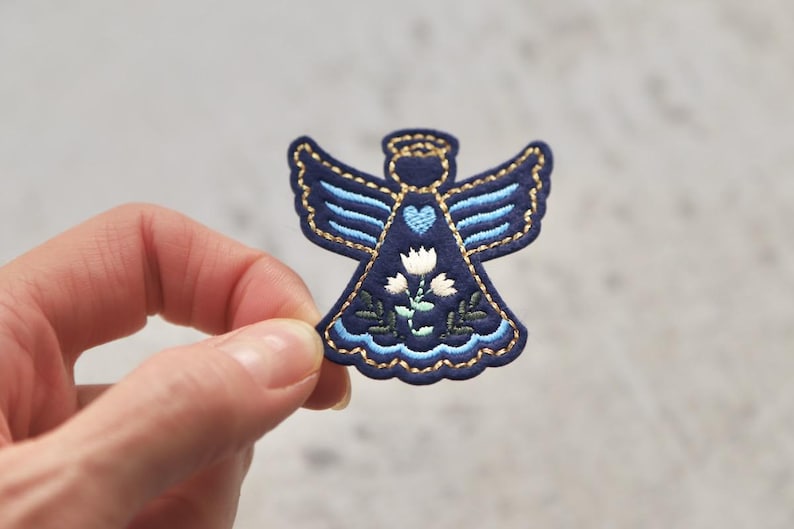 Peut inclure: Un ange en feutre bleu avec des d&eacute;tails brod&eacute;s, y compris un c&oelig;ur, des fleurs et des feuilles. L'ange a des fils dor&eacute;s qui d&eacute;limitent les ailes et la robe.