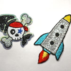 May include: Two iron-on patches, one with a silver glitter skull and crossbones with a red and blue pirate hat, and the other with a silver glitter rocket ship with a blue and yellow flame.