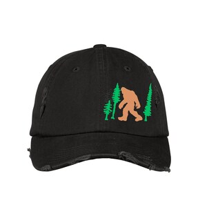Peut inclure: Casquette de baseball noire usée avec une silhouette de Bigfoot brune marchant dans une forêt d'arbres verts.