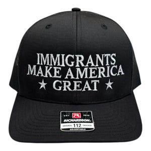 May include: Black trucker hat with white embroidered text reading "IMMIGRANTS MAKE AMERICA GREAT" with star accents. The hat features a mesh back and a curved brim. A Richardson brand tag is visible.