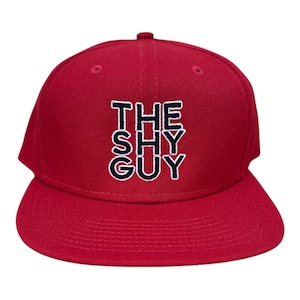 May include: A red baseball cap with a flat brim. The cap features the text "THE SHY GUY" embroidered in white with a navy blue outline. The hat is a classic six-panel design.