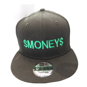 Puede incluir: Gorra de béisbol negra y verde con el texto "$MONEYS" bordado en la parte delantera. La gorra es una New Era 9FIFTY Snapback.
