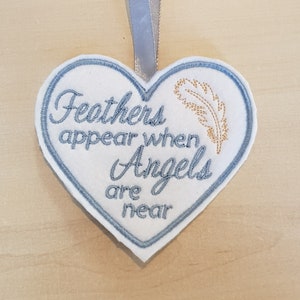 May include: White heart-shaped felt ornament with blue stitching and the text "Feathers appear when Angels are near" in blue embroidery. A gold feather is embroidered on the right side of the ornament.