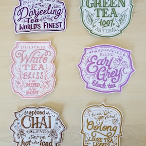 May include: Six embroidered tea bag labels featuring different tea varieties. The labels are white with colourful embroidery and text. The labels include Darjeeling Tea, White Tea, Earl Grey, Chai, Green Tea, and Oolong.