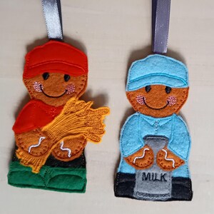May include: Two felt gingerbread person ornaments. One wears a red cap and shirt, holding wheat. The other wears a blue cap and shirt, holding a milk jug. Both have smiling faces and are trimmed with white stitching. Each has a ribbon for hanging.
