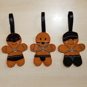 May include: Three gingerbread man ornaments with black leather straps and buckles. The gingerbread men are brown with pink cheeks and black eyes. They are hanging from black ribbon.