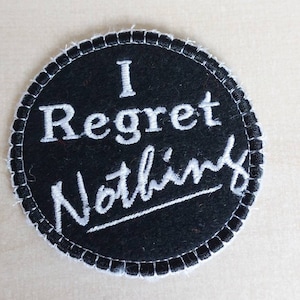 May include: Black and white embroidered patch with the text "I Regret Nothing" in white stitching.