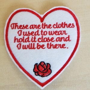 May include: Red heart-shaped patch with white fabric and red embroidered text that reads "These are the clothes I used to wear hold it close and I will be there." There is a red embroidered rose in the bottom centre of the patch.