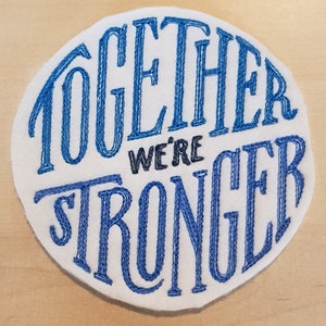 May include: White round patch with blue embroidered text that reads "Together We're Stronger".