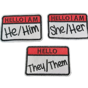 May include: Three white fabric name tags with black stitching and red lettering. The top two tags read "HELLO I AM" and have the pronouns "He/Him" and "She/Her" written in black. The bottom tag reads "HELLO" and has the pronouns "They/Them" written in black.