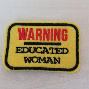 May include: Yellow felt patch with black stitching and the text "WARNING EDUCATED WOMAN" in black and red lettering.