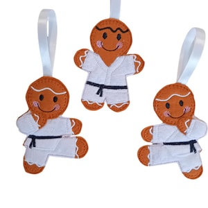 May include: Three gingerbread person ornaments wearing white karate uniforms with black belts. Each ornament has a ribbon for hanging.