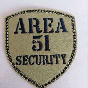 May include: A tan-coloured patch with a black embroidered border. The patch features the text "AREA 51 SECURITY" in black embroidery.