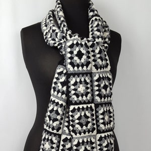May include: A black and white crocheted scarf with a square pattern and fringe. The scarf is draped over a black mannequin. The scarf's design features a repeating pattern of black, white, and gray squares, creating a textured look. The fringe at the ends adds a decorative element.