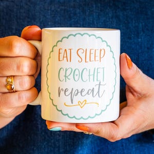 May include: White ceramic mug with the text "EAT SLEEP CROCHET repeat" in orange, teal, and grey, surrounded by a teal scalloped border. The mug is held by a person wearing a denim apron.