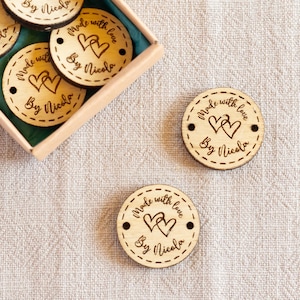May include: Circular wooden tags with the inscription "Made with love By Nicola" and heart designs. Some tags are in a small box with a green interior. The tags are light brown with dark brown text and illustrations.