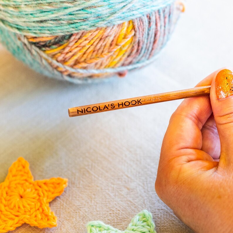 Puede incluir: Un ganchillo de madera con la inscripci&oacute;n "NICOLA'S HOOK" se sostiene cerca de una madeja de hilo colorido. El hilo es una mezcla de azul, verde, naranja y amarillo. Estrellas de ganchillo en naranja y verde en primer plano.