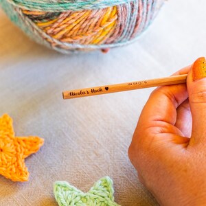 Puede incluir: Un ganchillo de madera con el texto "Nicola's Hook" y un s&iacute;mbolo de coraz&oacute;n. El ganchillo es de 5,0 mm. Una madeja de hilo multicolor est&aacute; en el fondo, junto con dos estrellas de ganchillo.