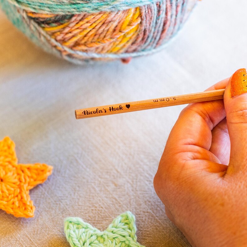 Puede incluir: Un ganchillo de madera con el texto "Nicola's Hook" y un s&iacute;mbolo de coraz&oacute;n. El ganchillo es de 5,0 mm. Una madeja de hilo multicolor est&aacute; en el fondo, junto con dos estrellas de ganchillo.