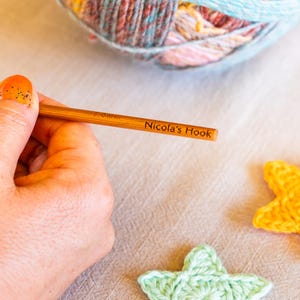 Puede incluir: Un ganchillo de madera con el texto "Nicola's Hook" se sostiene en una mano. El ganchillo est&aacute; junto a una madeja de hilo colorido y dos formas de estrella de ganchillo, una amarilla y otra verde.