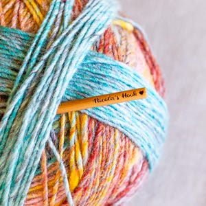 Puede incluir: Una madeja de hilo colorido con tonos de azul, naranja, amarillo y rojo. Un ganchillo de madera est&aacute; insertado en el hilo, con el texto "Nicola's Hook" grabado.