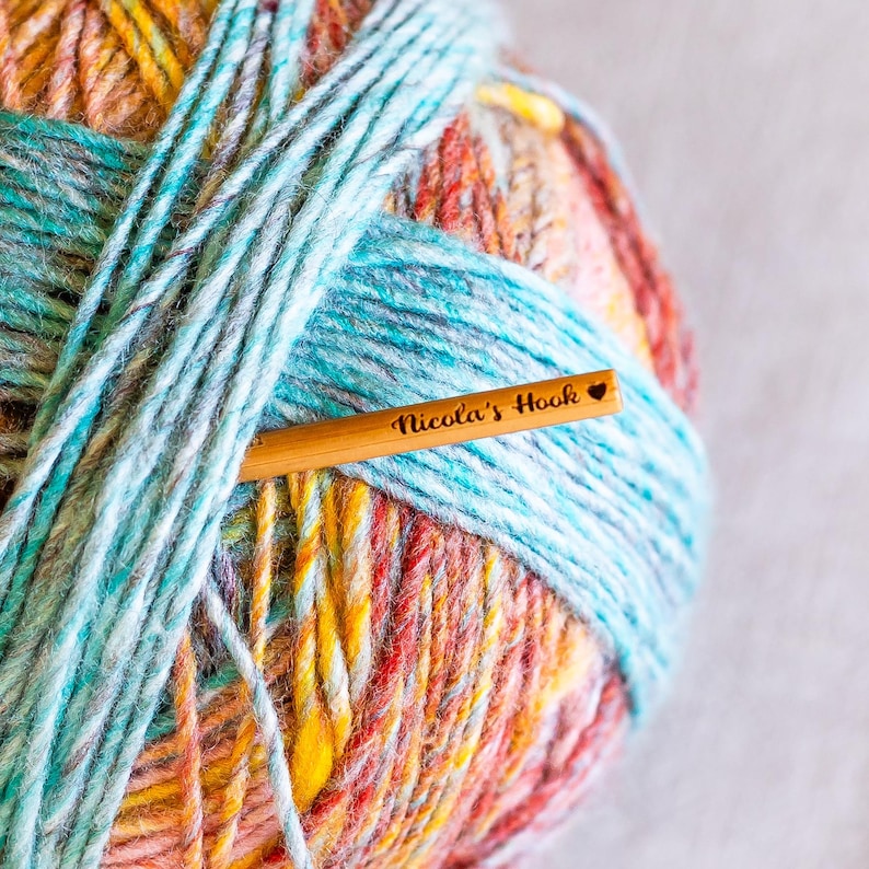 Puede incluir: Una madeja de hilo colorido con tonos de azul, naranja, amarillo y rojo. Un ganchillo de madera est&aacute; insertado en el hilo, con el texto "Nicola's Hook" grabado.