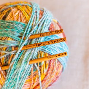 Puede incluir: Primer plano de una bola de lana multicolor con tres ganchillos de madera. La lana es una mezcla de amarillo, naranja, azul y verde. Los ganchillos llevan la inscripci&oacute;n "Nicola's Hook" y "5.0mm NICOLA'S HOOK".