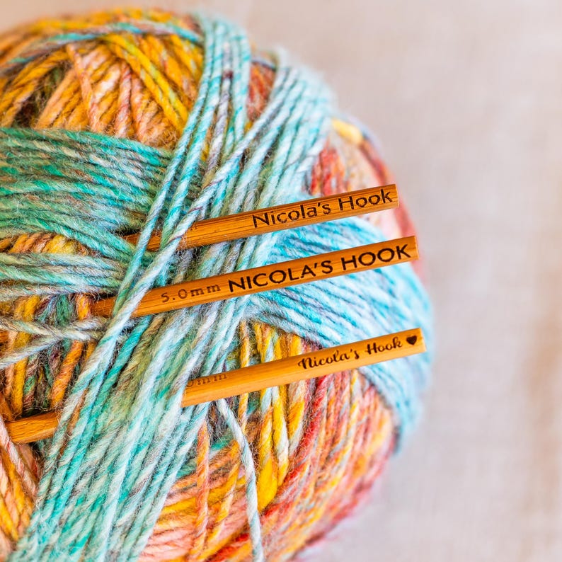 Puede incluir: Primer plano de una bola de lana multicolor con tres ganchillos de madera. La lana es una mezcla de amarillo, naranja, azul y verde. Los ganchillos llevan la inscripci&oacute;n "Nicola's Hook" y "5.0mm NICOLA'S HOOK".