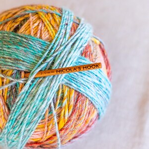 Puede incluir: Una madeja de hilo colorido en tonos amarillos, naranjas y turquesas, con un ganchillo de madera insertado. El ganchillo est&aacute; etiquetado como "5.0mm NICOLA'S HOOK". El hilo parece suave y texturizado, ideal para proyectos de tejido o ganchillo.