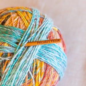Puede incluir: Una colorida madeja de lana con tonos amarillos, naranjas, azules y verdes. Un ganchillo de madera est&aacute; insertado en la lana, con el texto "Nicola's Hook" grabado. La lana parece suave y texturizada, ideal para proyectos de tejido o ganchillo.