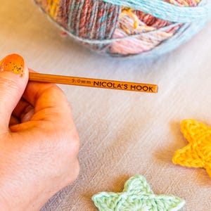 Puede incluir: Un ganchillo de madera con la inscripci&oacute;n "5.0mm NICOLA'S HOOK" se sostiene cerca de una madeja de hilo multicolor. Estrellas de ganchillo en amarillo y verde claro est&aacute;n en primer plano. El hilo es una mezcla de azul, rosa y marr&oacute;n.