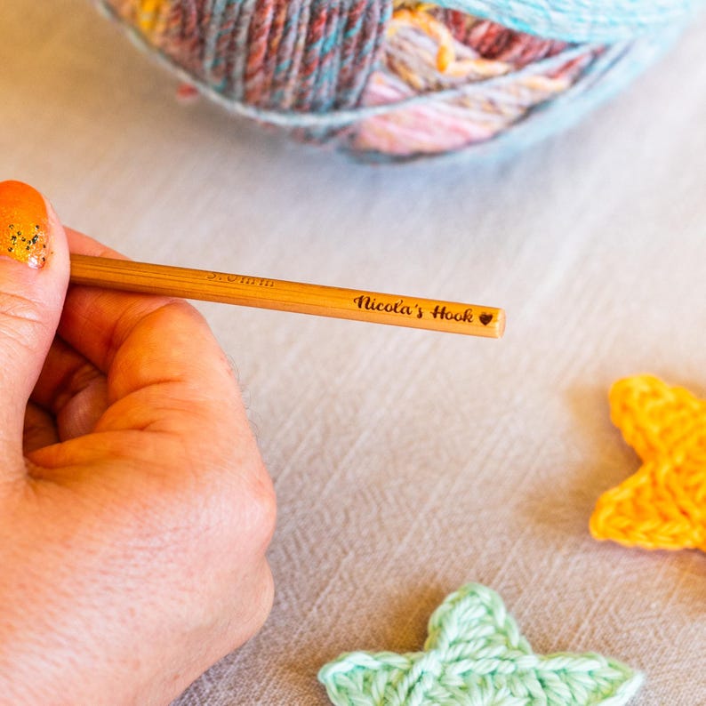 Puede incluir: Un ganchillo de madera con el texto "Nicola's Hook" y un s&iacute;mbolo de coraz&oacute;n. El ganchillo se sostiene en una mano, con una madeja de lana y dos formas de estrella de ganchillo en el fondo. El ganchillo es de 5,0 mm.
