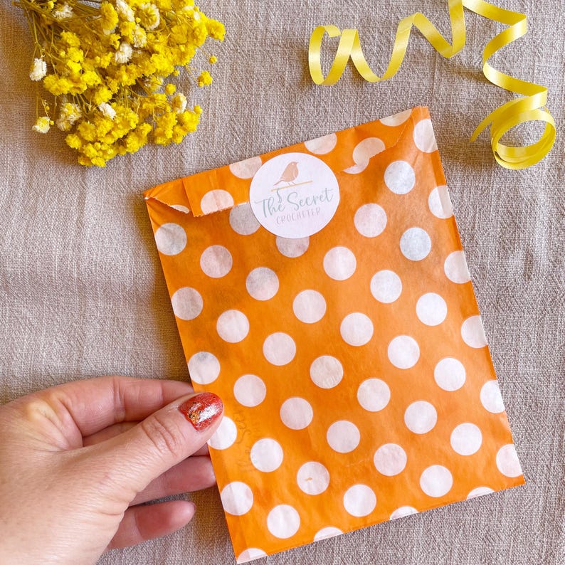 Puede incluir: Un paquete naranja con lunares blancos con una pegatina circular blanca que dice "The Secret Crocheter" es sostenido por una mano. Flores amarillas y una cinta amarilla est&aacute;n en el fondo. El paquete est&aacute; sellado.