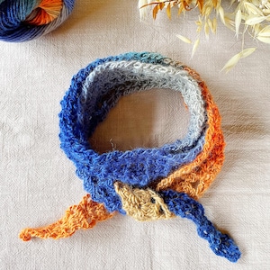 May include: A handmade crochet scarf in shades of blue, gray, and orange. The scarf is knotted at the center and has a triangular shape. A ball of yarn in similar colors is visible in the upper left corner. The scarf is made of soft yarn.